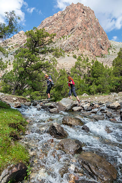 Two hikers crossing fast flowing river People going across mountain creek with fast streaming water jumping on the rocks green meadow and forest along river mountain landscape and sky on background ford crossing stock pictures, royalty-free photos & images