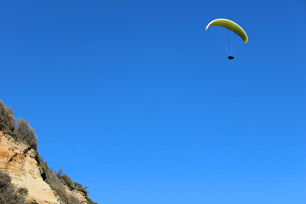 Photograph showing an unrecognisable paraglider flying high over the coastline, above eroding cliffs made of sandstone.