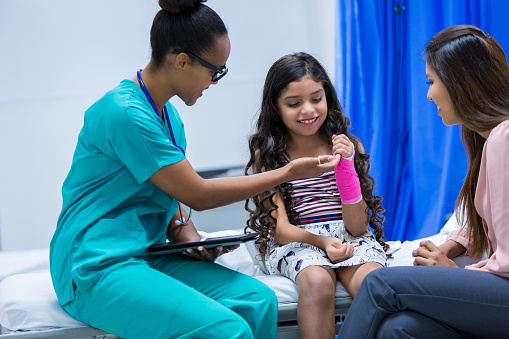 Young adult African American female doctor is examining injured arm of pediatric patient. Patient is elementary age Hispanic little girl who is wearing a pink cast on her wrist. Mother is comforting patient while doctor examines injury in hospital emergency room.
