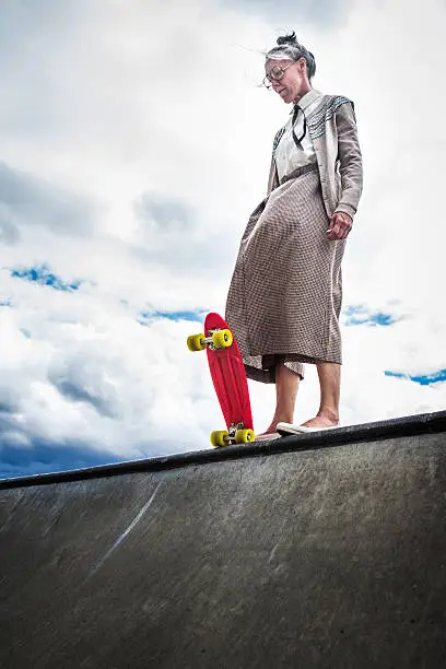 A senior elderly woman attempting skateboarding stands at the edge of a halfpipe debating whether to drop in.   A great additional exercise and balance for her healthy lifestyle.