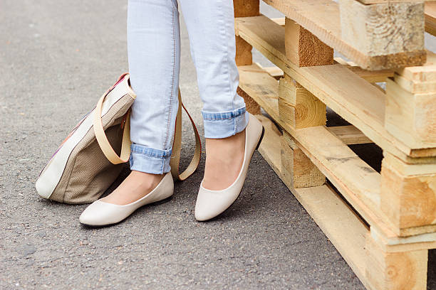 Woman's legs in jeans and flat shoes Woman's legs in jeans and white ballet flat shoes with beige bag, standing near wooden palettes flat shoe photos stock pictures, royalty-free photos & images
