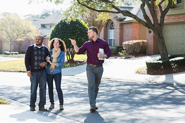 Black couple walking on residential street with agent An African American couple walking on a residential street, with a young man holding a folder.  He is a real estate agent, pointing at a house, talking. real estate agent male stock pictures, royalty-free photos & images