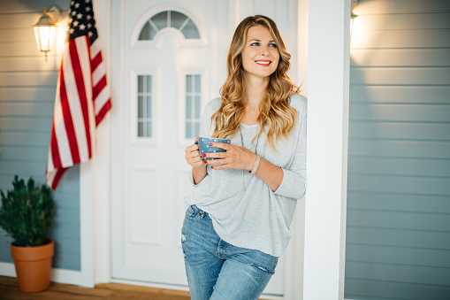 Woman standing on a porch of her house, American flag in background. Holding coffee mug.