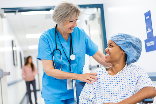 Senior adult Caucasian female nurse with gray hair is pushing patient in wheelchair. She is taking patient to recovery after outpatient surgical procedure. Patient is young adult African American woman, who is wearing a hospital gown and surgical cap. Women are smiling while talking to each other.