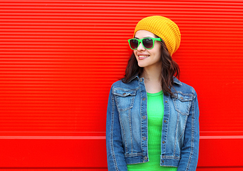 Fashion portrait of pretty young smiling woman wearing a sunglasses and colorful clothes against the red background