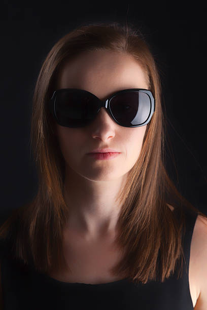 Cute brunette wearing a black dress, photographed on a dark background in dark sunglasses.