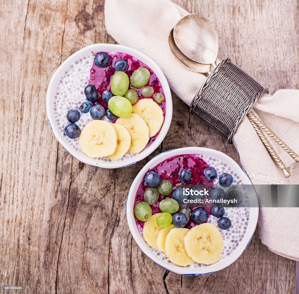 Chia Pudding for breakfast in bowls garnished with berry smoothies Chia Pudding for breakfast in bowls garnished with berry smoothies, grapes, petals of almond and blueberry in two portions on a wooden background with a napkin and spoon. Top view. selective Focus 2015 Stock Photo