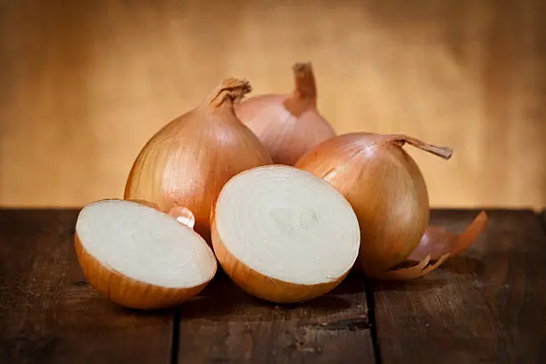 Still life of three gold onions on a wooden table, the one on the foreground it is cut in halves. Predominant color is brown. DSRL studio photo taken with Canon EOS 5D Mk II and Canon EF 70-200mm f/2.8L IS II USM Telephoto Zoom Lens