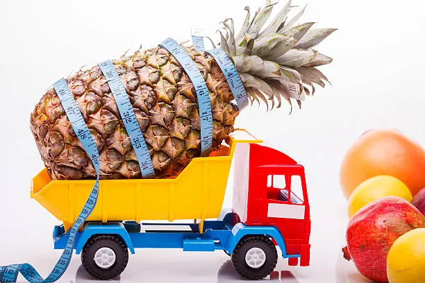 Toy lorry yellow blue red colors with pineapple fruit and measuring tape as diet symbol in basket with orange grapefruit pomegranate and lemon on white studio background closeup, horizontal picture