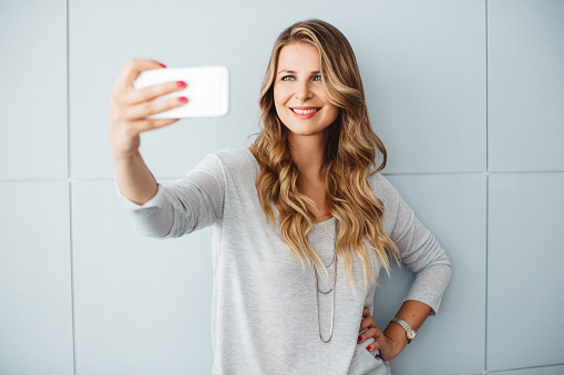 Beautiful mid-age woman standing by blue wall in office or home, using smart phone to take selfies. Casual clothes, caucasian. Long blond hair.