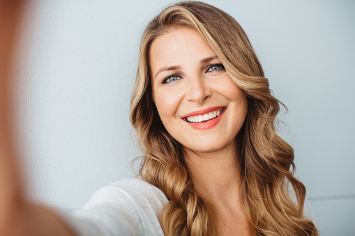 Beautiful mid-age woman standing by blue wall in office or home, using smart phone to take selfies. Casual clothes, caucasian. Long blond hair.