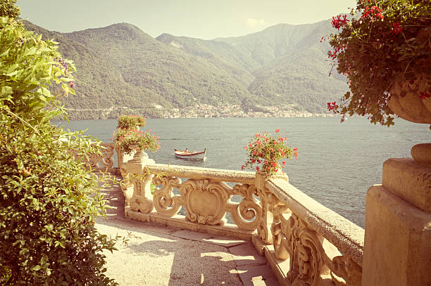 Lake Como, Italy. An old stone balcony, stone pots with flowers in the foreground, against beautiful lake, mountain and rock view. Heavy processing for retro look.