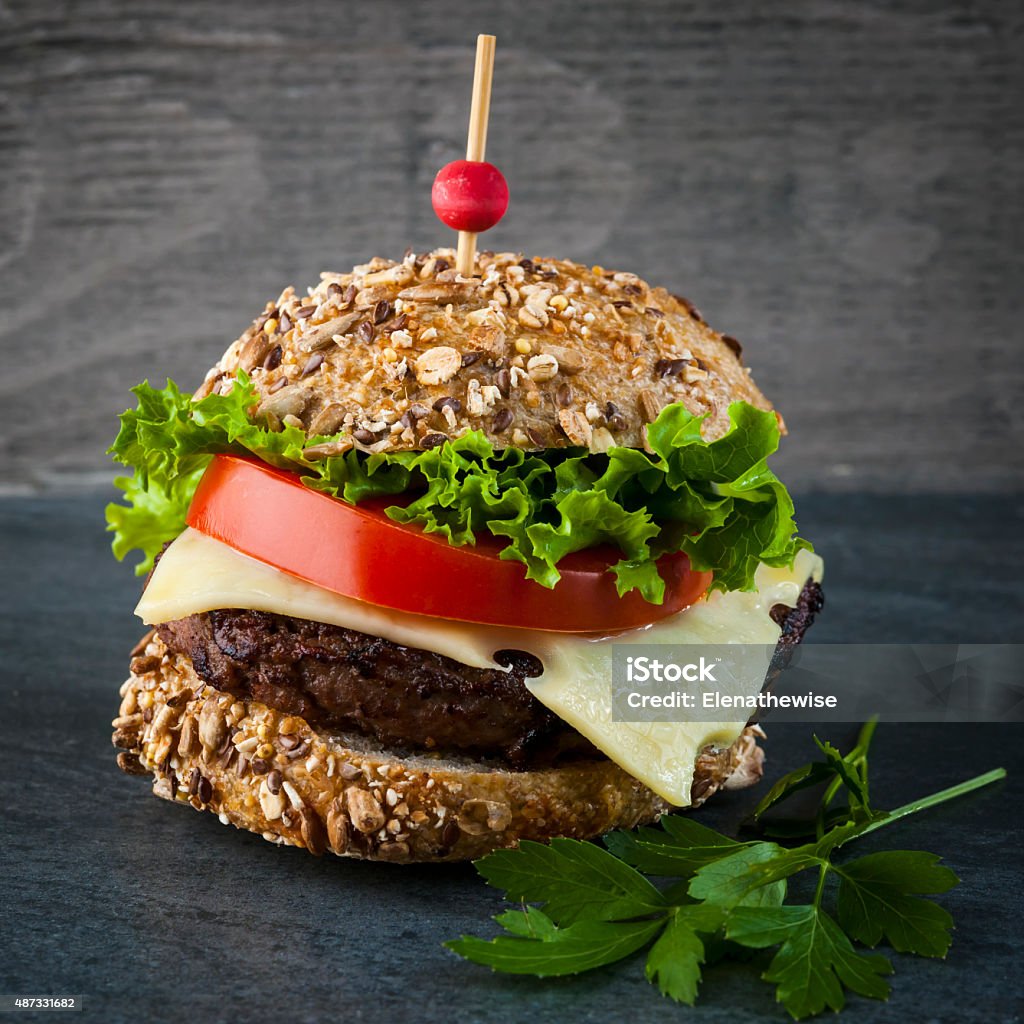 Gourmet hamburgers Gourmet hamburger with swiss cheese and fresh vegetables on multigrain bun over dark background, square format 2015 Stock Photo