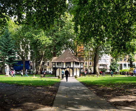 London, England - September 3, 2015: Lunchtime in Soho Square, Central London, with people walking, sitting and eating lunch. Soho Square was created in the late 17th century and was once a highly fashionable place to live, but the old buildings now house a variety of businesses, many related to media industries. The ancient-looking half-timbered building in the centre of the square is a gardeners’ hut.