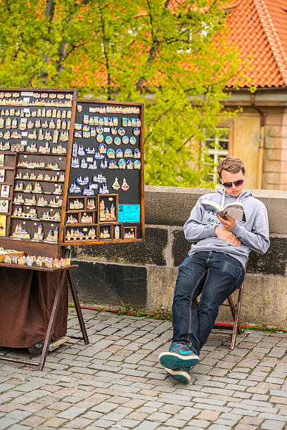 Prague, Czech Republic - May 3, 2015: Young man selling souvenirs on Charles Bridge, Prague, waiting for customers and reading a book