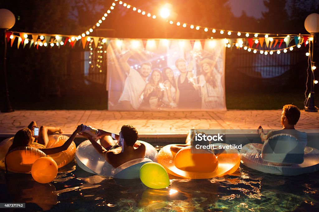 Pool movie night party. Young friends having pool movie night party. Floating on the pool on inflated mattresses and watching movie on improvised screen. Backyard decorated with festive string lights. Night time.  Movie Theater Stock Photo