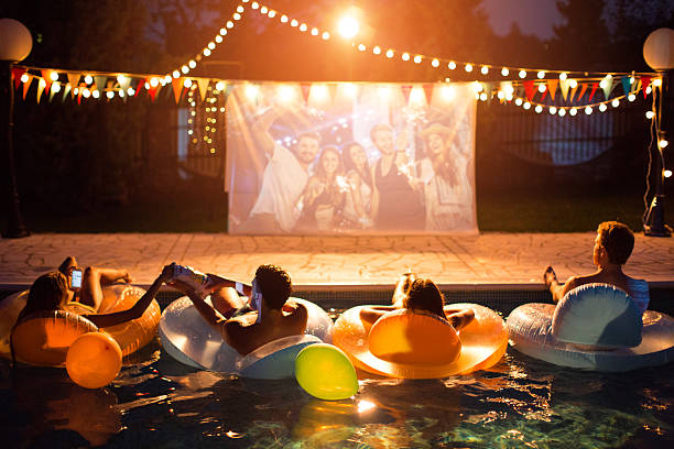 Pool movie night party. Young friends having pool movie night party. Floating on the pool on inflated mattresses and watching movie on improvised screen. Backyard decorated with festive string lights. Night time.  movie theater photos stock pictures, royalty-free photos & images