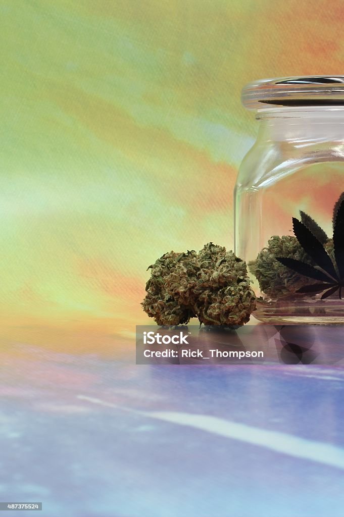 Medical marijuana in a jar with rainbow background image right Buds of marijuana displayed in a glass jar bearing an etching of a marijuana leaf. The background is a hippie tie dye colorful setting with blue and yellow as the dominant colors. There is ample room for logos or ad copy in this vertical image, useful for any legalization or medical marijuana purpose. The imagery is confined to the center-right area, exposing a greater portion of the image for text or copy. The colors are bright and the image crisp.  2015 Stock Photo
