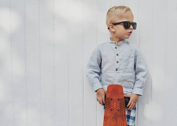 Cute little guy in big trendy sunglasses that he has borrowed from his mother or father posing with his skateboard against a white wooden wall with copyspace