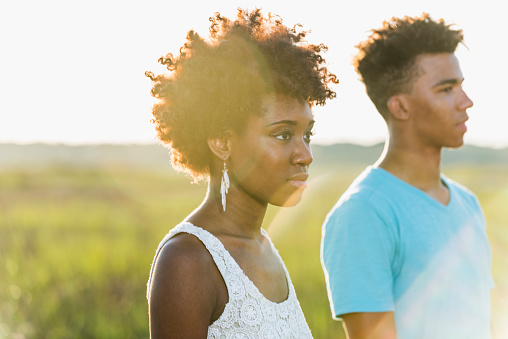 Portrait of a young African American couple with serious expressions, standing in sunlight in a field, looking away.  We see their head and shoulders.  The focus is on the woman.