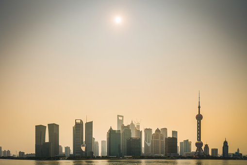 Golden sun shining through hazy skies silhouetting the futuristic skyline of Pudong with the iconic spire of the Oriental Pearl Tower and skyscrapers of the Lujiazui financial district above the Huangpu River, Shanghai, China. ProPhoto RGB profile for maximum color fidelity and gamut.