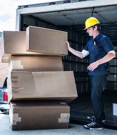 A careless manual worker young man is stacking heavy merchandise boxes haphazardly onto his hand truck dolly on the loading dock at the back of his open delivery truck. Most of the cardboard cartons already appear damaged. Slight motion blur on the carton he is holding.
