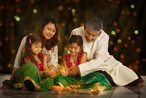 Indian family in traditional sari lighting oil lamp and celebrating Diwali or deepavali, fesitval of lights at home. Little girl hands holding oil lamp indoors.