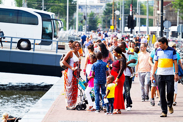 Amsterdam, The Netherlands - August 9, 2015: Summertime shot of black people and families with children and baby buggies at Oosterdokskade. At leftv side is Open Havenfront. In background are many people and a bus crossing a bridge.