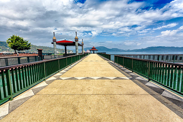 Small pedestrian bridge over river Wang in Lampang. View from southern riverside. A few thai people are crossing the bridge. Bridge is also open for motorcycles. Above bridge is multi colored penant decor on lines. and ropes In right background are buildings at street Thipawan Alley. In sky dark storm clouds are approaching