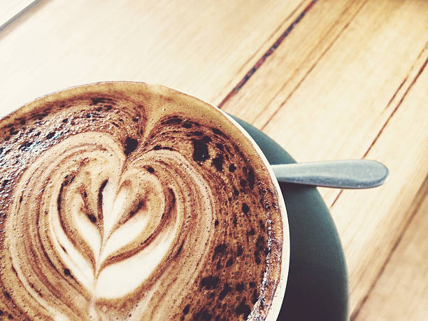 A cappuccino on a black saucer with a spoon on a bench in a cafe made by a barista. there is a heart shaped pattern on top of the coffee/crema.