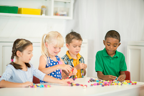 A multi-ethnic group of elementary age children are sitting at a desk stringing together beads for an arts and crafts project.
