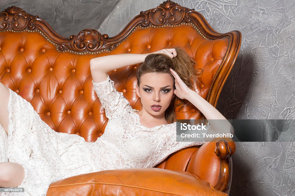 Lying woman in dress Beautiful confident young stylish woman in white lace dress with blonde hair and bright makeup looking forward lying on brown leather royal sofa on grey wall background, horizontal picture 2015 Stock Photo