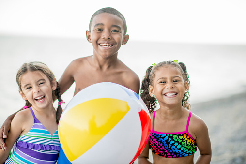 A multi-ethnic group of elementary age children are outside standing together holding a beach ball at the beach. They are smiling and looking at the camera.
