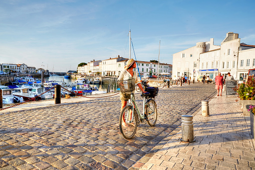 St. Martin-De-Re, France - June 25, 2015: Boats in harbour at St Martin-De-Re. Woman cycling on cobblestoned harbour at St Martin-De-Re. Other people are pictured walking in the background