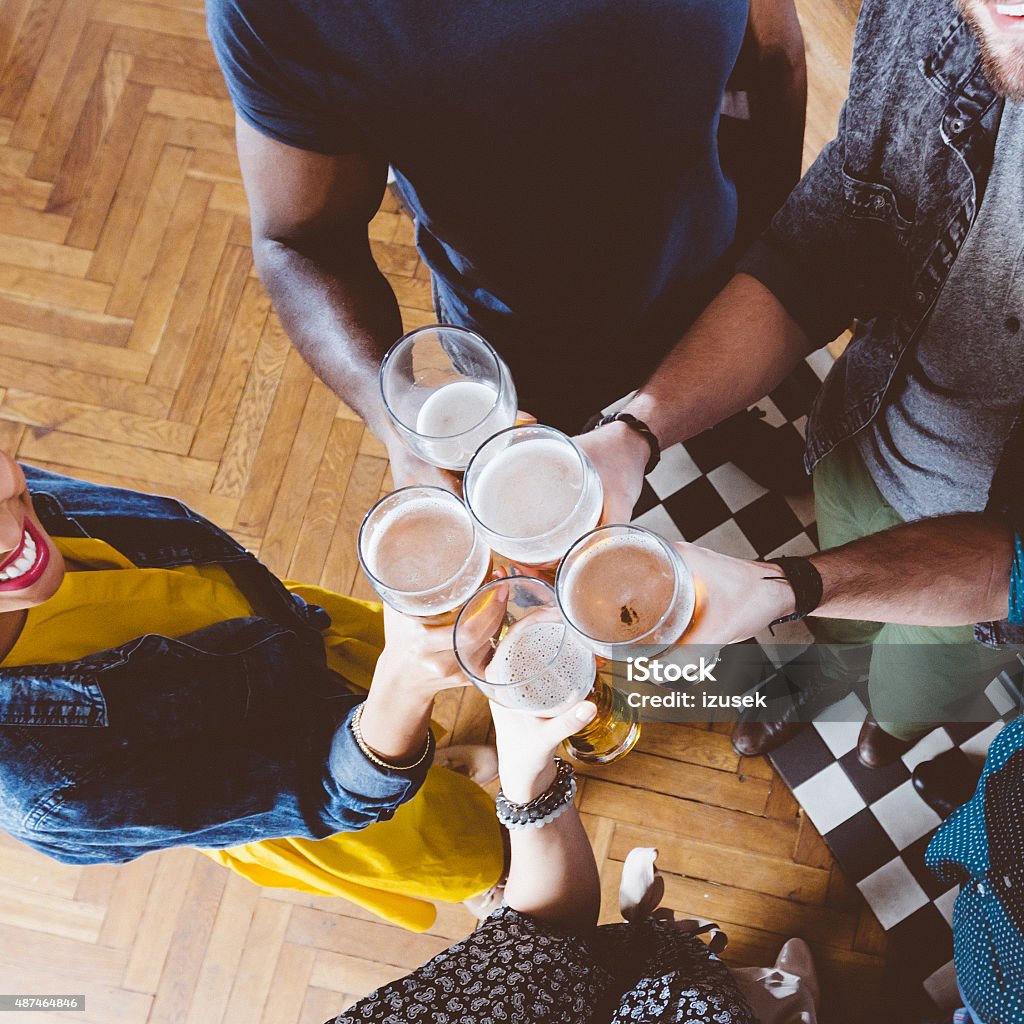 Friends toasting with beer in a pub Multi ethnic group of friends toasting with beer glasses. High angle view, close up of hands. Unrecognizable people.  Beer - Alcohol Stock Photo