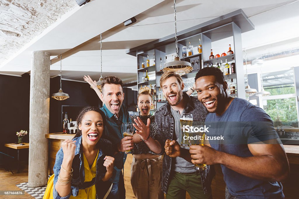 Friends cheering with beer in a pub Multi ethnic group of happy friends having party in a pub. Cheering with beer glasses.  Sport Stock Photo