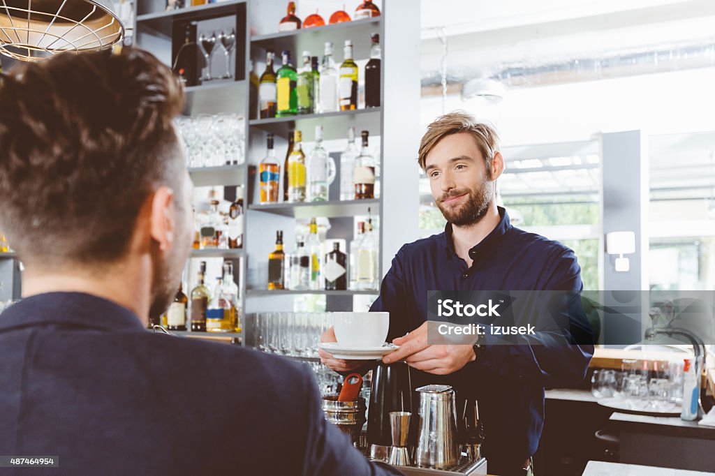 Barman serving coffee in a pub In the pub. Smiling bearded barman serving coffee to a businessman. 2015 Stock Photo