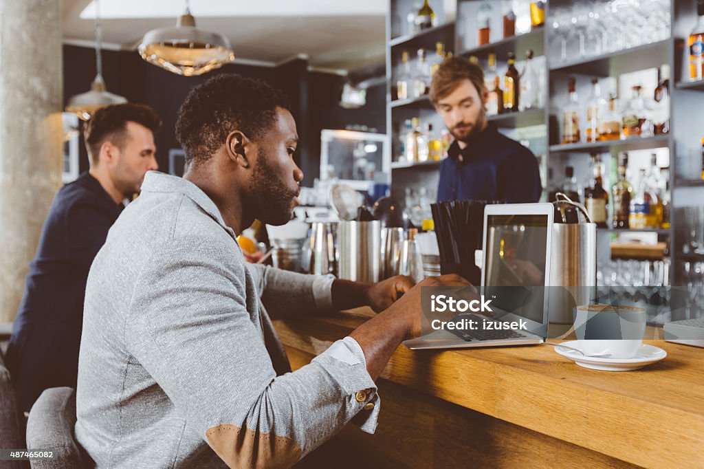 Afro american man using laptop in a pub Afro american man wearing jacket sitting by the counter in the pub and using laptop. Bearded barman in the background.  Incidental People Stock Photo
