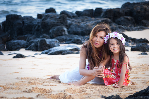 A beautiful Asian/Hawaiian mother and daughter are wearing a Lei around the neck and head. The woman is wearing a long white dress and the little girl is wearing an orange dress..They are sitting on the sand on Poipu beach, at a resort in Kauai. They are sitting closely together and appear very relaxed and happy to be on vacation.