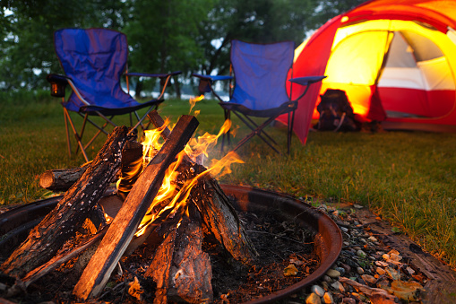 Horizontal view of evening campsite. A red tent set up on green grass, a fire pit burning a campfire in the foreground, chairs, cooler, lamp and cooking equipment suggest relaxation in a natural night setting.