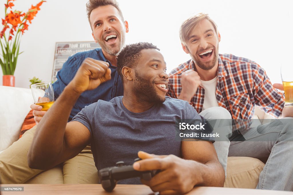 Three guys playing video games Multi ethnic - caucasian and afro american - friends sitting on sofa at home, playing video games and drinking beer.  Video Game Stock Photo