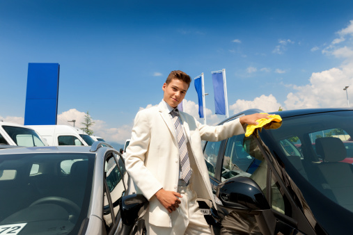 Used Car Salesman shining a car with a yellow clothes, dressed in a white suit, looking at camera