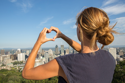 Young cheerful woman in Montreal, Canada making a heart shape with her hands. Point of view taken from the top of the Mont Royal park.