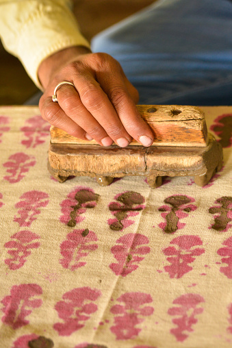 This is a vertical closeup image of a man's hand woodblock-printing on a fabric textile. He is demonstrating the ancient Indian textile art using mud as a resist. This was shot in a village in India.