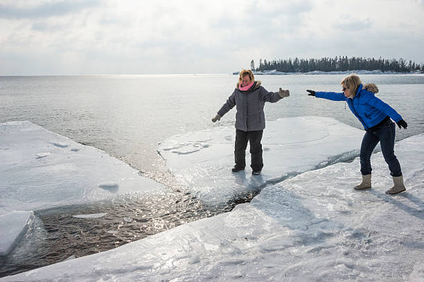 Caucasian adult woman reaches out to a blond woman balancing on an ice floe floating in Lake Superior in winter, Grand Marais, MN, USA