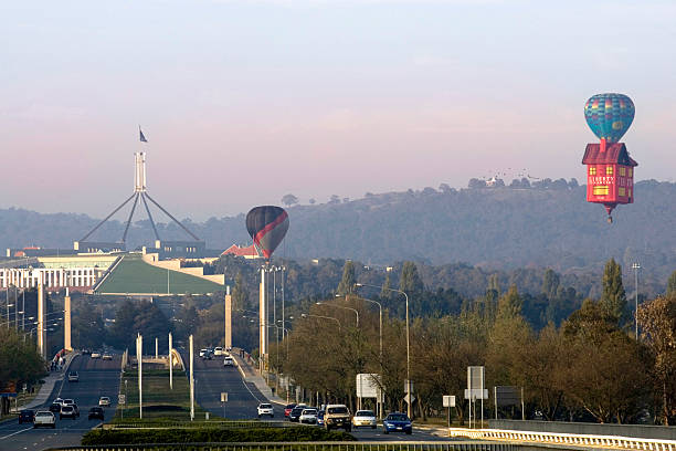 Canberra, Australia - April 14, 2007: Sunrise over main road in Canberra with hot air balloons and Parliament house in the distance, people riding in air balloon and people in cars driving around.