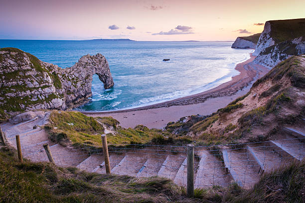 Path to ocean beach cove cliffs Durdle Door Dorset UK Warm glow of sunset light illuminating the steps down to the iconic rock arch of Durdle Door and the surf lapping the sheltered beach bay on the Jurassic Coast of Dorset, UK, a UNESCO World Heritage Site. ProPhoto RGB profile for maximum color fidelity and gamut. durdle door stock pictures, royalty-free photos & images