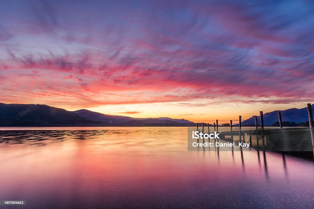 Stunning Vibrant Sunset With Jetty Pier In The Lake District. A photograph taken at the Ashness Gate jetty/pier at Derwentwater in Keswick, Lake District, UK. The photograph features a vibrant orange and purple sunset, the beautiful Cumbrian mountains in the background and the jetty to the right of the image. English Lake District Stock Photo
