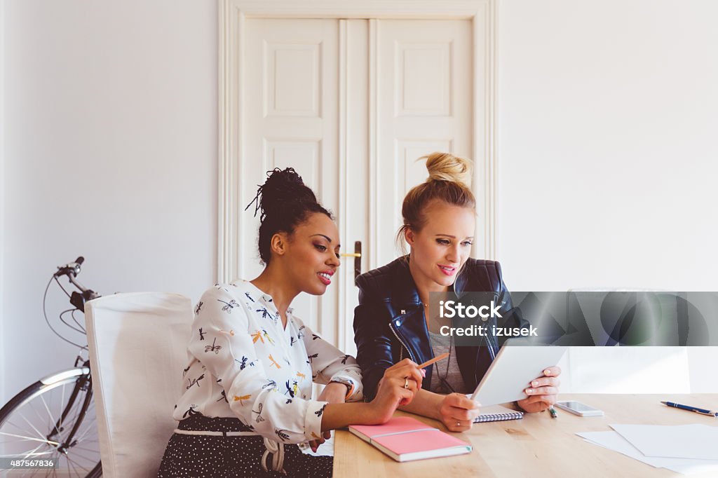 Two women working on digital tablet in an office Start-up agency. Two women - caucasian and afro american - sitting at the table in an office and using a digital tablet together.   Social Media Stock Photo