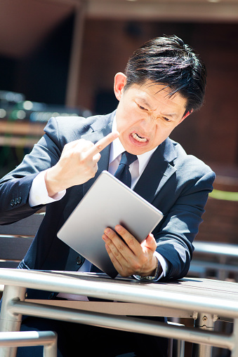 Furious Japanese businessman gives the finger to his tablet during a particularly frustating video conference. Photographed outdoors at a Shijuku cafe in Tokyo, Japan.